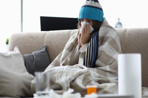 sick person sitting on couch with blankets and a hat on